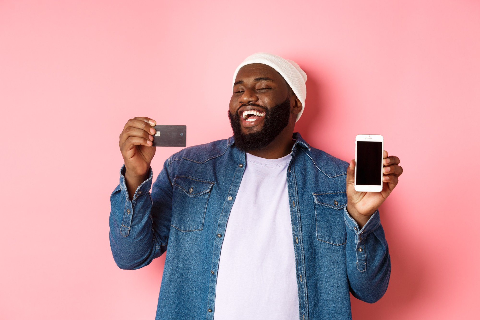 online-shopping-happy-african-american-man-beanie-laughing-showing-credit-card-mobile-phone-screen-standing-pink-background