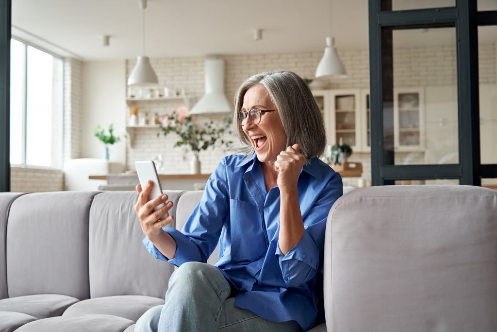Excited mature old woman winner reading message on smartphone at home.