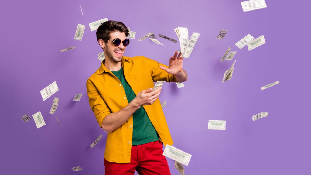 Photo of cheerful excited white trendy man throwing cash away, showing his wealthiness smiling toothily in red pants isolated violet color background