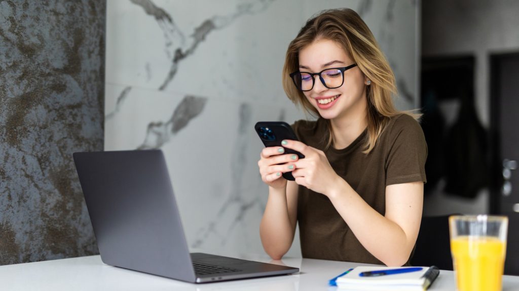 pretty woman with eyeglasses sits office using laptop and smartphone cellphone