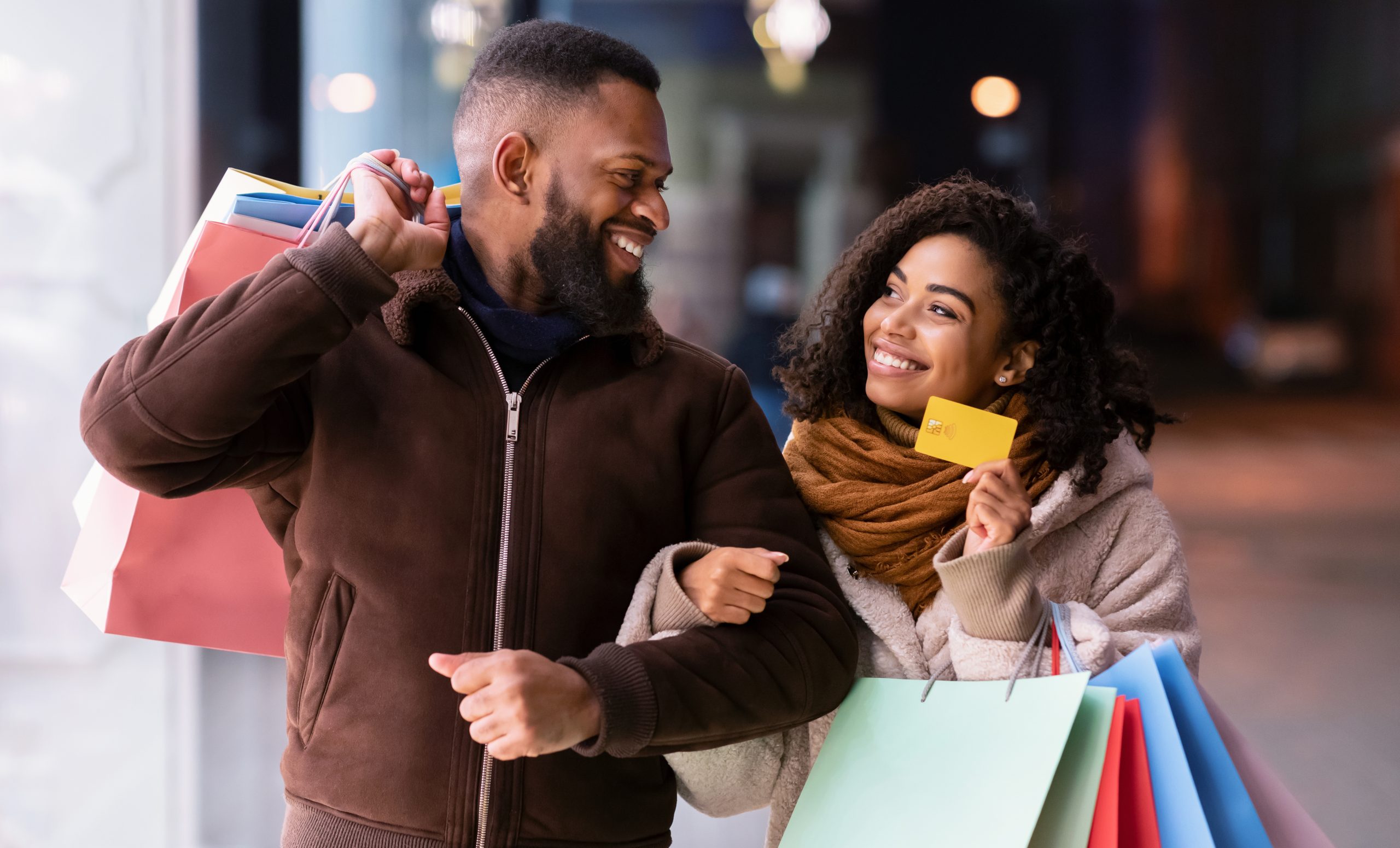 Let's Buy Something. Portrait of smiling black woman showing debit credit card to man, holding shopping bags, walking outside in the city near mall in the evening, blurred background