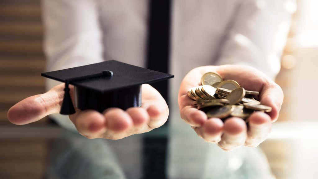 Human Hand Holding Black Graduation Hat And Golden Coins Over Desk