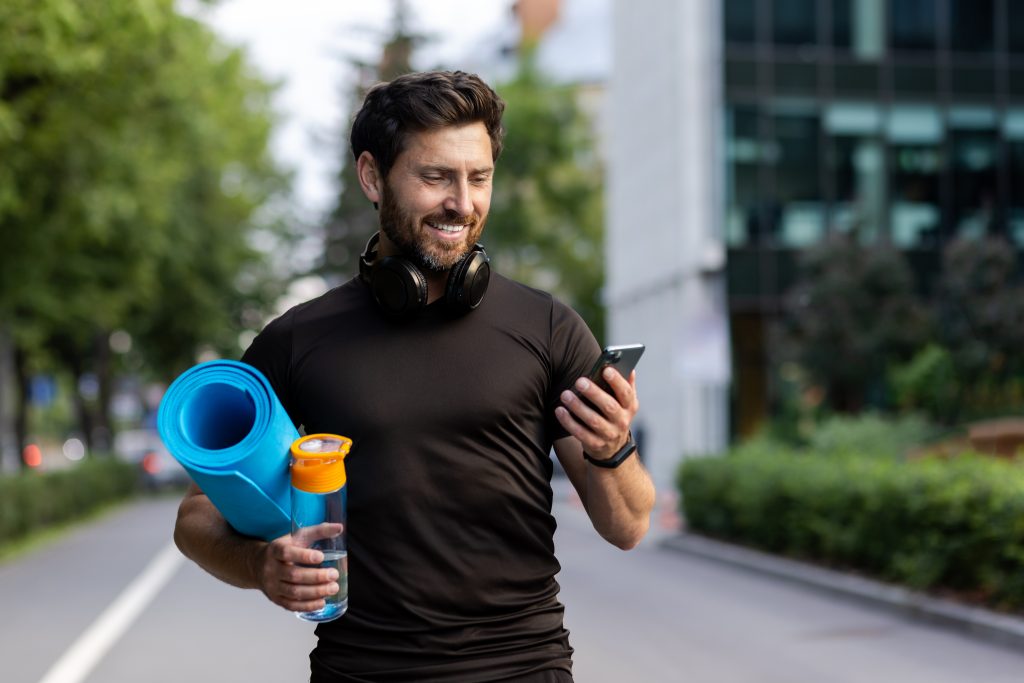 Portrait of mature successful adult athlete, bearded man going to workout, coach holding fitness mat and phone, browsing online app for joint physical training